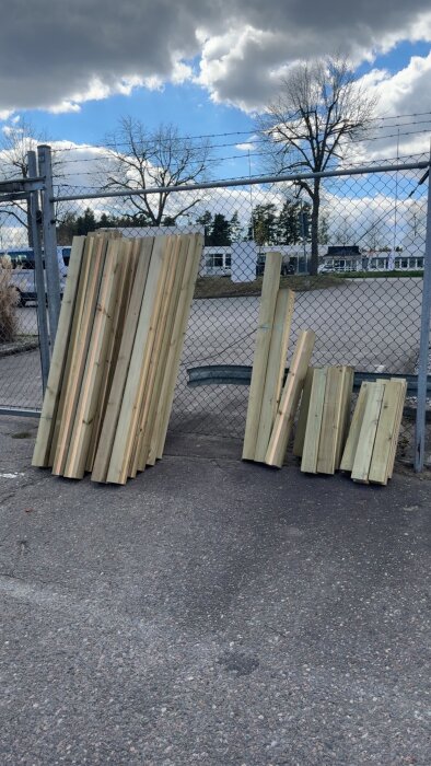 Brädor som står lutade mot ett staket, synliga moln och träd i bakgrunden. Fotot är taget utomhus på asfalt.