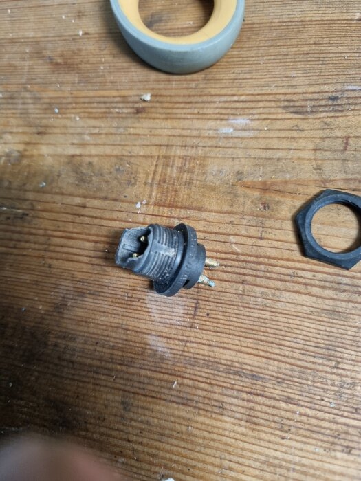 Trasigt ladduttag med lösa stift från en Husqvarna 305 ligger på en träyta bredvid en rulle tejp och en ringformad komponent.