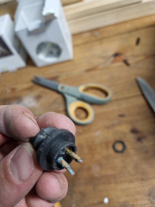 Hand håller en liten svart kontakt med två stift, omgiven av en suddig bakgrund med en sax och lådor på ett träbord.