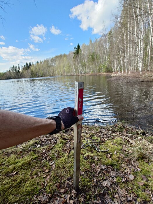 En hand håller en mätpinne vid en sjö med skog i bakgrunden och blå himmel med vita moln ovanför.