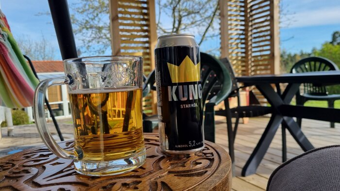 En burk med starköl och ett glas öl står på ett utsmyckat bord på en altan med trämöbler i bakgrunden.