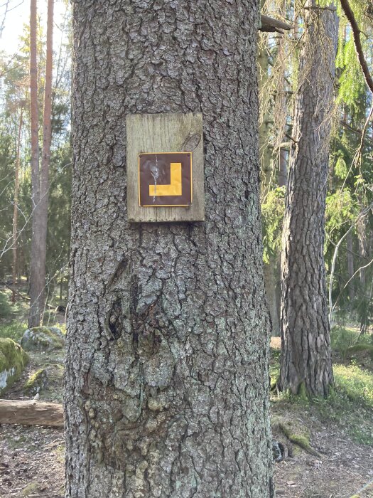 Trädstam i skog med träskylt, gul-orange L-formad symbol på brun bakgrund. Solbelysta träd i bakgrunden.