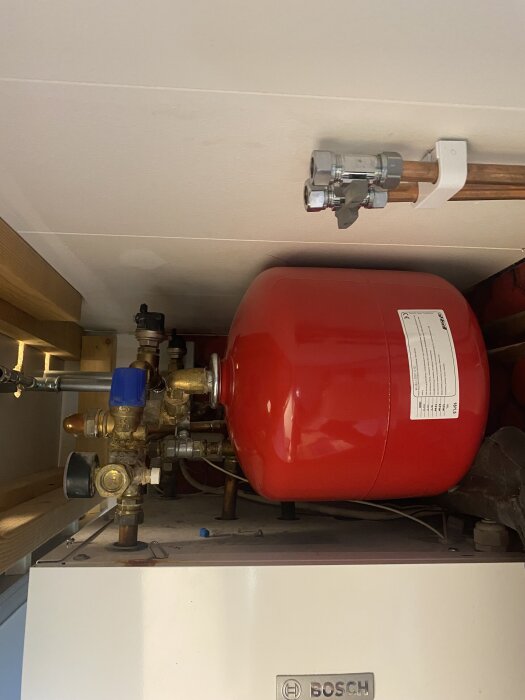 Röd expansionskärl vid en Bosch bergvärmepump med kopparrör och ventil inuti ett hus.