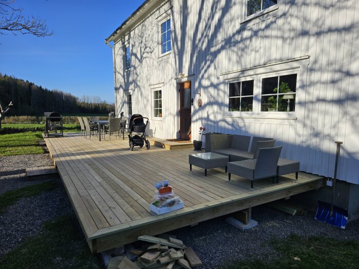 Nybyggd altan med trädäck, möbler, barnvagn och solbelyst vit husvägg i bakgrunden. Uttag för stolpar syns i hörnen.