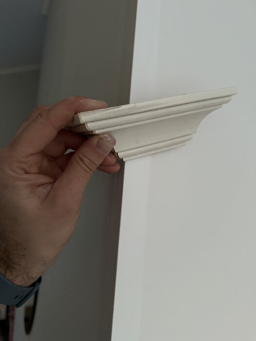 En hånd holder en hvitmalt list mot en vegg for å illustrere vinkeljustering ved montering.