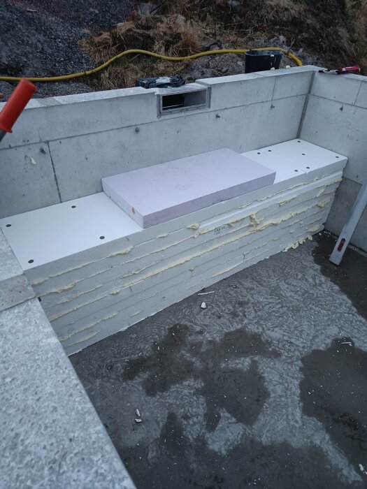 Bänk under konstruktion i större bad, gjord av vit cellplast med dräneringshål, armering och fogband, täckt av plast; omgiven av betonggolv.