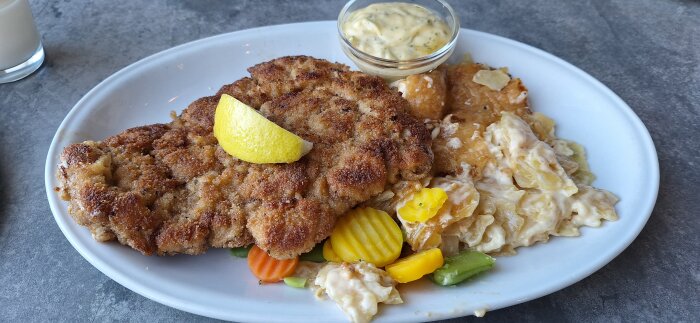 Tallrik med schnitzel toppad med citron, potatisgratÀng, morötter, bönor och bearnaisesÄs i skÄl.