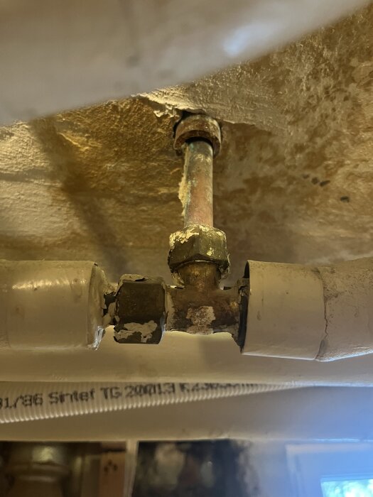 Kopparrör med T-koppling under ett tak, med anslutning till två vita PEX-rör, visar äldre installation för vattenanslutning.