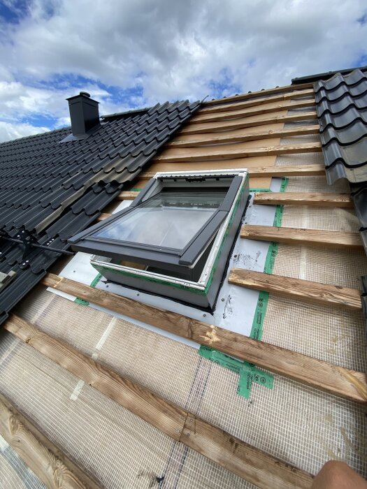 Takfönster under reparation visar delvis avslöjat tak med träreglar, underlagspapp och blottlagda fästen på ett hus med grå takpannor.
