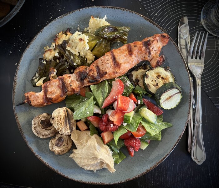 Grillat kycklingspett med champinjoner, squash, sallad, hummus, och ugnsgrillade padrones med ost fyllning på en tallrik.