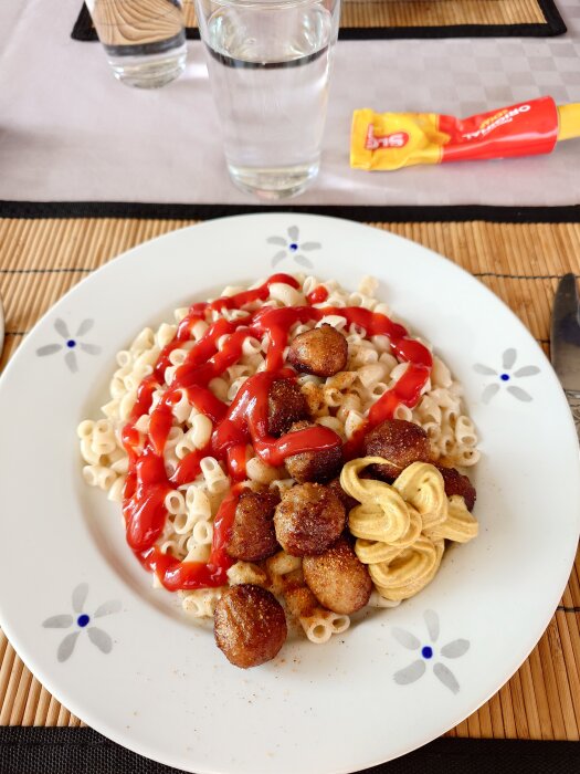 Stuvade makaroner med köttbullar, ketchup och senap pÄ en tallrik, glas vatten i bakgrunden.