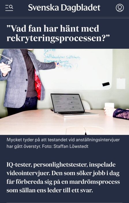 En person i kavaj pekar på en whiteboard med text, bredvid står en bärbar dator och en kopp på ett bord.