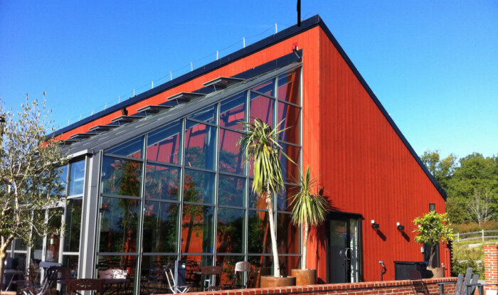 Rött modernt hus med snedtak och stora glasfönster, omgiven av träd och utemöbler.