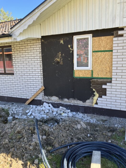 Fasad av hus under renovering med nytt fönster, borttagen mexisten, och utsprida tegelstenar på marken.