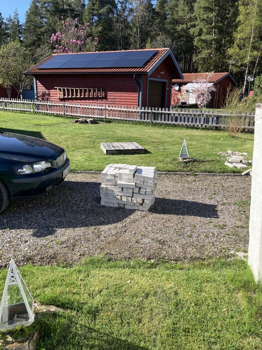 Hög med mexistenar på en grusgång, solpaneler på rött hus i bakgrunden, bil synlig till vänster, soligt väder och grönt gräs.