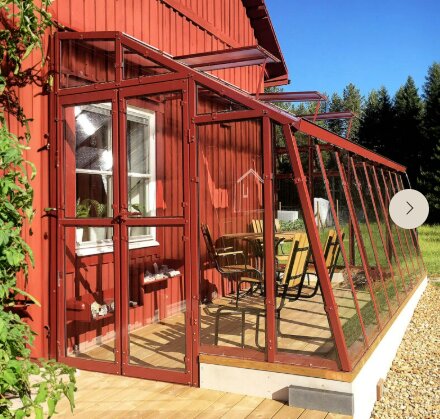 Rött hus med en tillbyggnad av ett väggväxthus i glas, som inkluderar stolar och ett bord. Omgiven av grönska och stenbelagd gång.