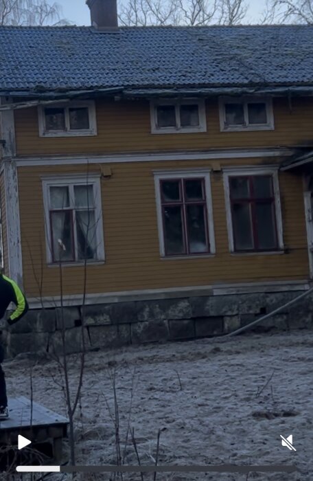 Gult trähus med blåa takpannor och vitmålade fönster, synlig grund av stora stenar; marken runt huset är frostbelagd.