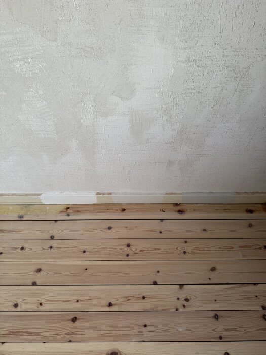 Oputsad vägg med ojämn gipstextur och synligt trä/randigt golv i förgrunden. Test av olika tekniker för att jämna ut och dammbinda ytan.