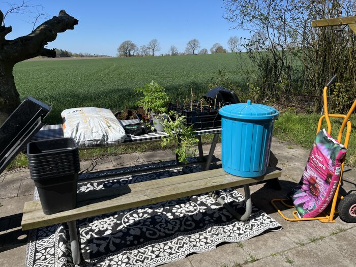 Planteringsplats med picknickbänk, krukor, planteringsjord och en blå soptunna, med utsikt över gröna fält och träd.
