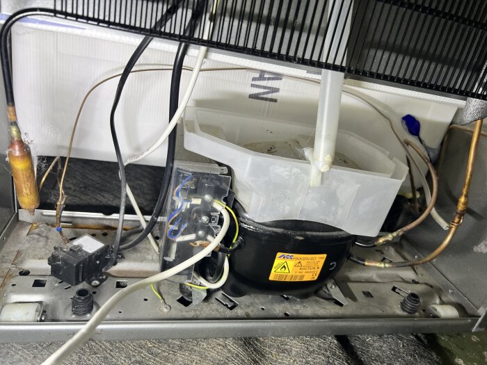 Kylskåpets baksida med kompressor, kablar och rör synliga. Kompressorn har en varningsetikett.