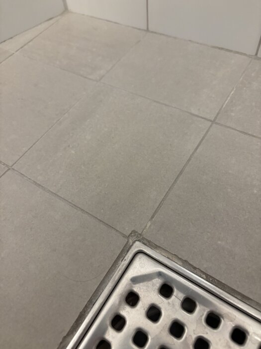 Grå kakelplattor på badrumsgolv med vit missfärgning på fogarna runt golvbrunnen.