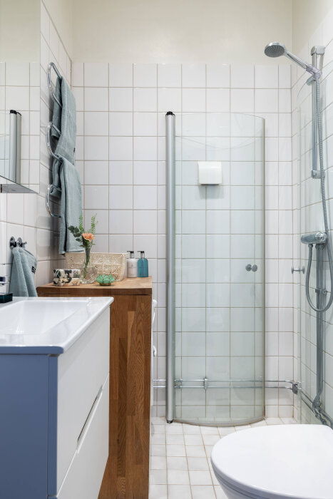 Modernt, kompakt badrum med vita kaklade väggar, glasdusch, vägghängd handdukstork, träskiva med handfat och dekorativa accessoarer.