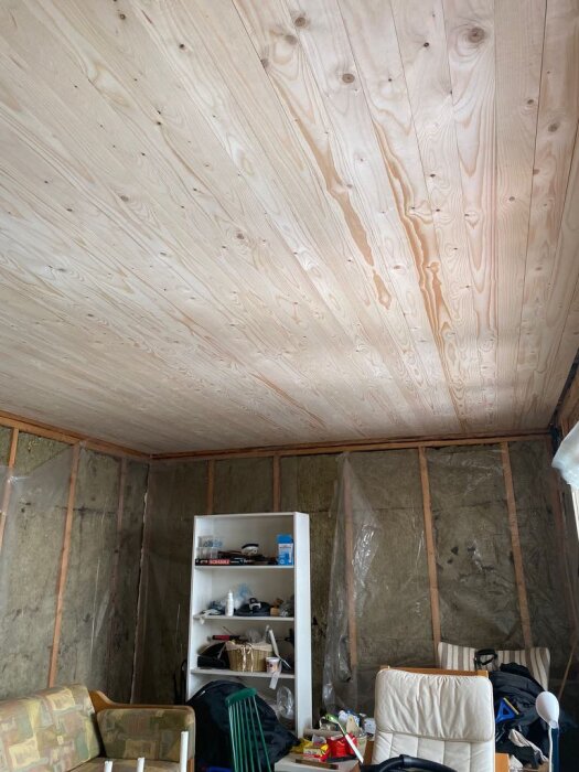 Innertak av råspont i en stuga under renovering, med isolerade väggar, hyllor och stolar nedanför.