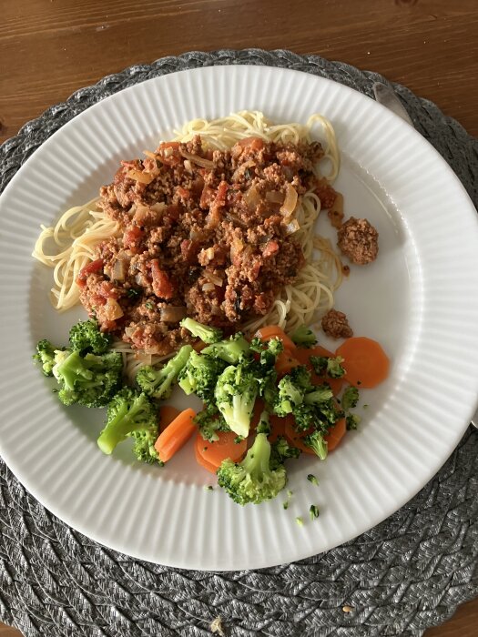 Pasta med köttfÀrssÄs toppad med chili och basilika pÄ en tallrik, serverad med broccoli och morotsskivor.