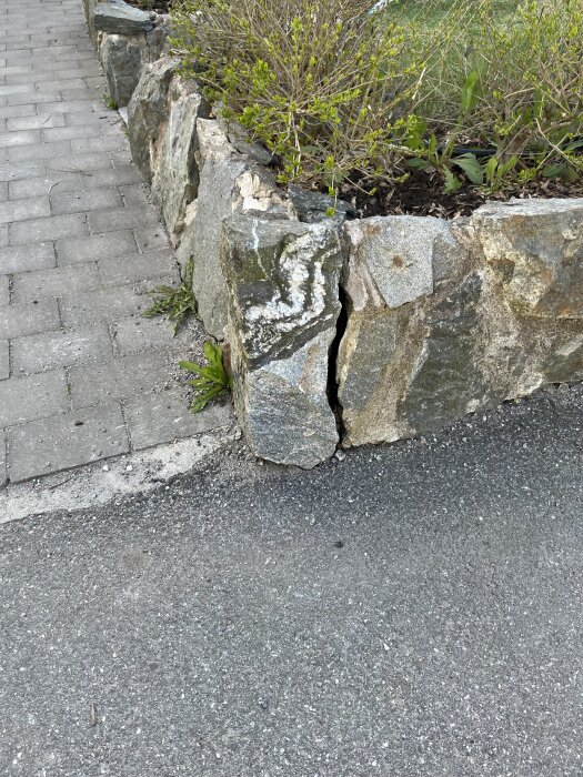 En skadad stenmurs hörnsten som har lossnat efter att ha blivit påkörd av en bil, bredvid en trottoar och växtlighet.