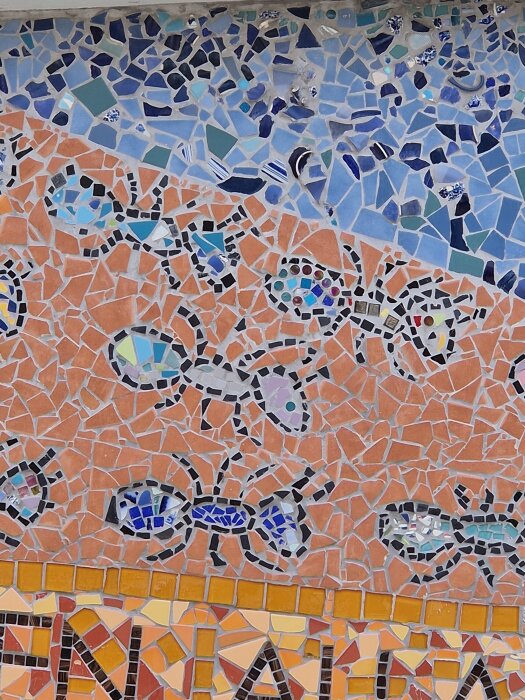 Mosaikvägg med färgglada myror i svart, blått och lila mot en bakgrund av orange och blåa stenar.