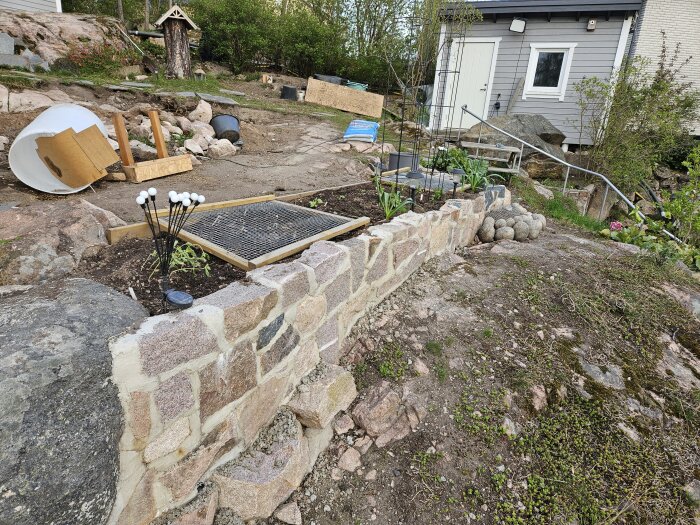 Altan med stenmur, jord och växter på ojämn mark, trappa i bakgrunden, föremål och vegetation runt området.