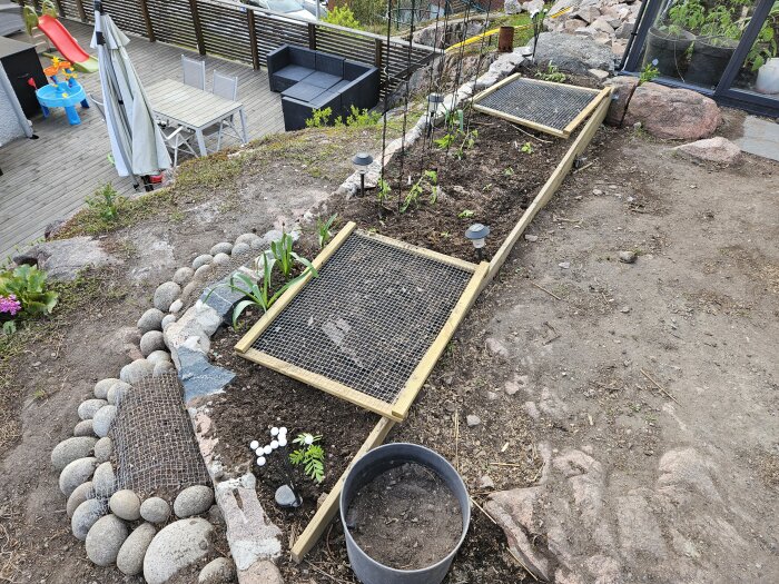 Planteringslådor på altan med jord och nät. Omgiven av stenar och växter. Terrass i bakgrunden med utemöbler och leksaker.
