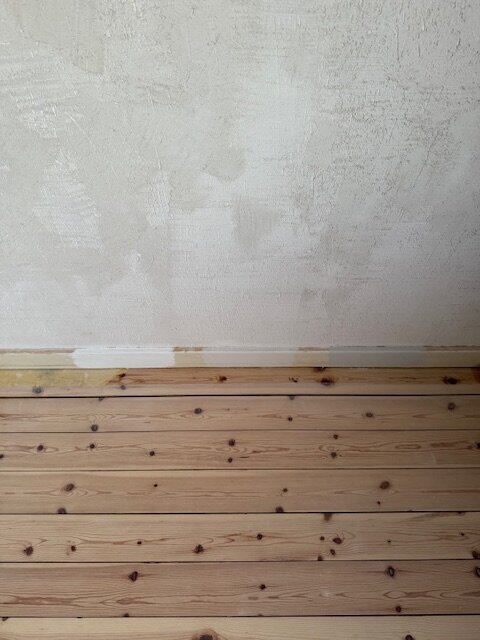 Bild av en vägg under renovering med ojämn gipsputs och ett trägolv framför. Väggen ser skrovlig ut med fläckar av olika texturer.