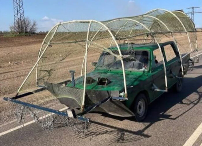 Grön bil ombyggd med metallram och nät, liknande ett improviserat stridsfordon, på väg på landsbygden.