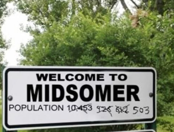 Skylt med texten "Welcome to Midsomer" och en överstruken befolkningssiffra som ändras från 10453 till 503, omgiven av grönska.