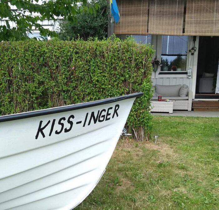 Vit plasteka med namnet "Kiss-Inger" målat på fören, ligger på gräsmatta framför ett hus med buskar runt.