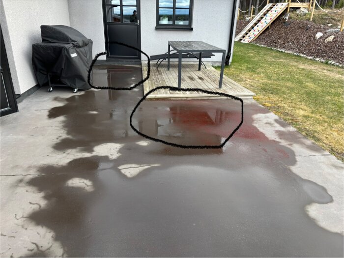 Betonguteplats med vattenpölar, markerade cirklar kring pölarna. Uteplatsen angränsar till huset med ett bord och en grill. Gräs och trätrappa i bakgrunden.