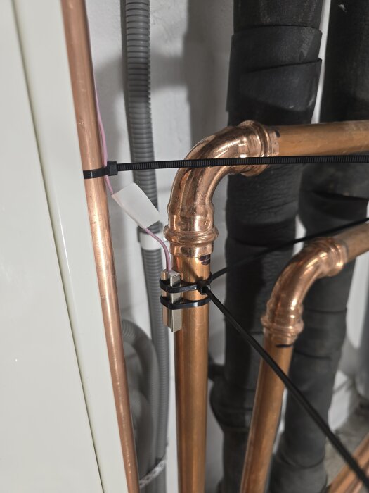 Kopparrör med termistor fäst med buntband vid rörkorsning, omgivna av isoleringsmaterial och elektriska ledningar i teknikskåp.