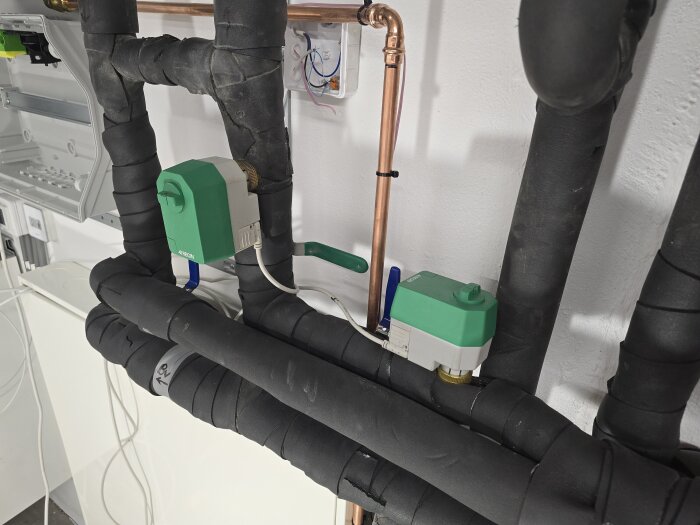Isolerade rör med gröna reglermotorer installerade vid ett elskåp, del av ett kyl- och värmesystem för golvkylanläggning.