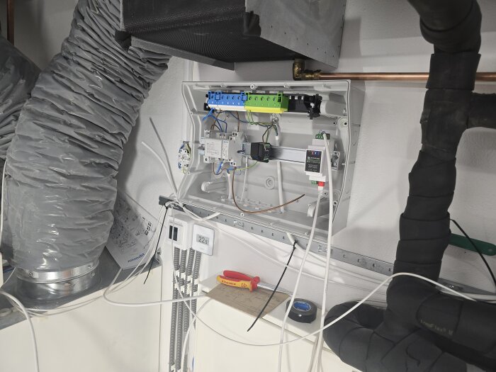 Öppet elskåp med kablar, kopplingar och komponenter för kylsystem, luftslangar, och en Shelly-modul monterad på väggen i tekniskt rum.