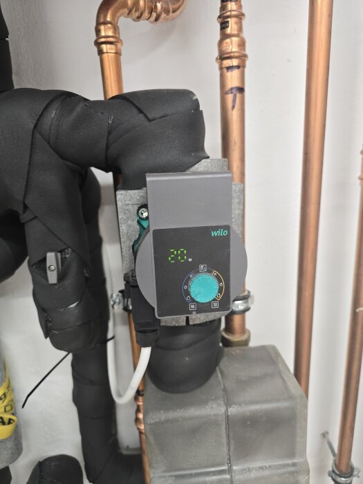 En Wilo-cirkulationspump installerad bland kopparrör och omgiven av isolering, med en inställning om 20 watt synlig på displayen.