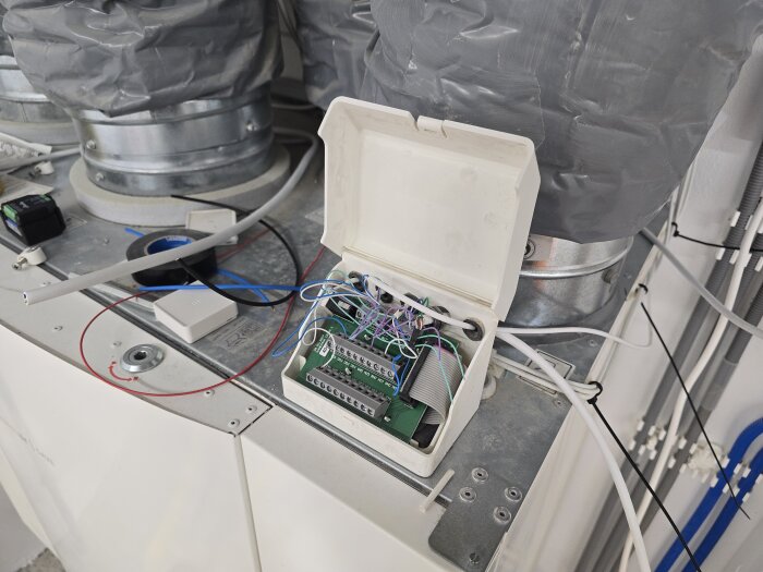 Öppet elskåp med kopplingsplintar och kablar, placerat ovanpå en metallisk yta, omgiven av rör och ledningar.