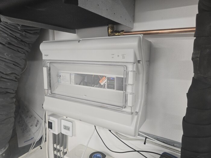 Elskåp med styrsystem för golvkyla och luftkyla installerat på en vägg, omgivande rör och isolerade kanaler synliga.