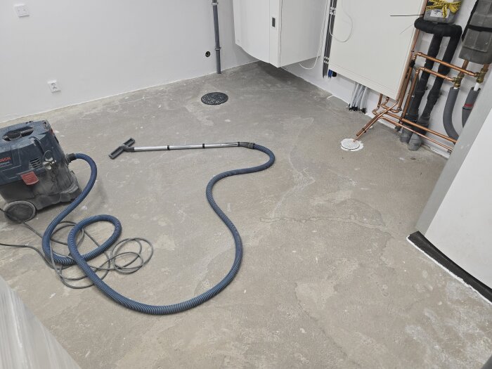 Industridammsugare och kopparrör i en renoverad byggarbetsplats med synligt betonggolv, förberett för plattsättning.