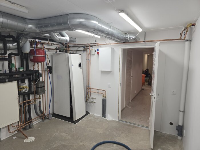 Bild av ett teknikrum med synliga rörledningar, ventilationssystem och värmepumpsutrustning. Golvet är ofärdigt och rummet är under renovering.