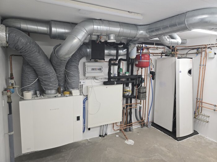 Elskåp med rör och ventilationssystem installerade mot en vit vägg i en källare. Inkommande vatten, FTX, kylreglering och värmepump syns.