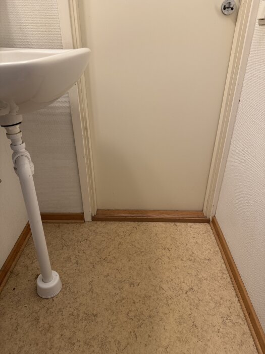 Toalett med dörr vid ena kanten och handfat. Golvet har ett mönster och dörrkarmen är i trä.