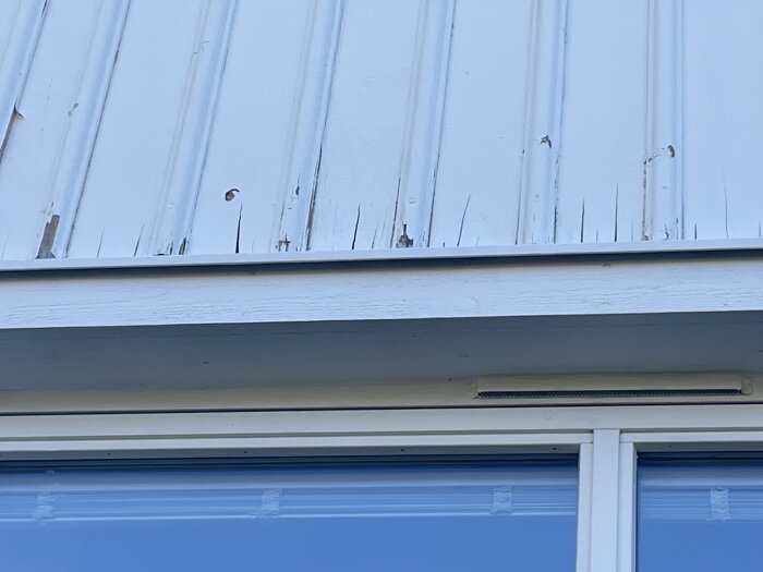 Sprucken och slitna fasadbrädor ovanför ett fönster på en byggnad.