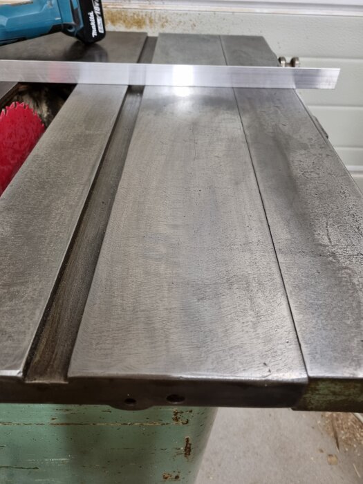 Metallbord slipat med sandpapper och abralon, sett ovanifrån. Röd klinga synlig under bordet och en metalldel ligger tvärs över ytan.