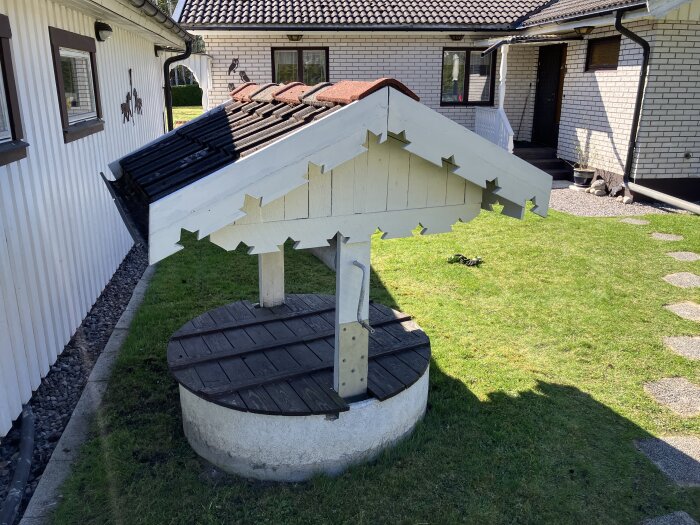 Hissanordning vid brunn i trädgård, vit träkonstruktion med takpannor ovanpå. I bakgrunden syns ett hus och gräsmatta.